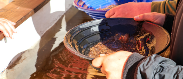 Två händer som håller i en vaskpanna vid rinnande vatten. I pannan syns sand som kanske kan innehålla guld.