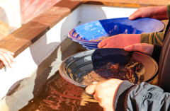 Två händer som håller i en vaskpanna vid rinnande vatten. I pannan syns sand som kanske kan innehålla guld.