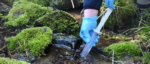 Bilden visar två händer som arbetar med att provta grundvatten med hjälp av en spruta.