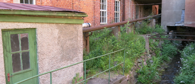 På bilden syns en röd tegelvägg från det tidigare pappersbruket i Fengersfors i bakgrunden. Framför det står en mindre kontorsbyggnad och från den röda tegelbyggnaden förbi det mindre huset rinner en konstgjord bäck.