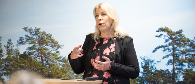Anette Madsen står och pratar framför en fondvägg med tallar. Anette Madsen står och pratar framför en fondvägg med tallar.