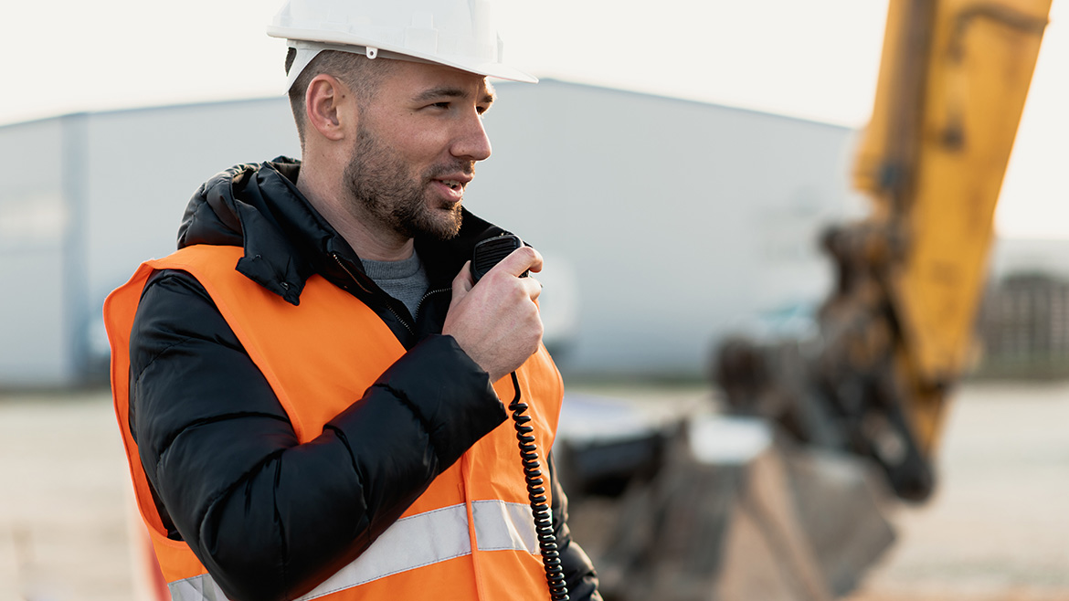 En man med varselväst och hjälm pratar i en walkie-talkie. Foto.