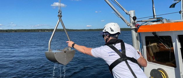 Båt, hav och person som tar sedimentprover med hjälp av skopliknande verktyg.
