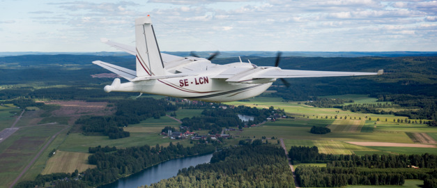 Flygplan som flyger över landskap med skog och vatten.