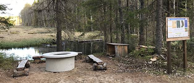 Tre stycken träbänkar runt en eldplats i betong. I bakgrunden syns vatten och en skog. Foto.