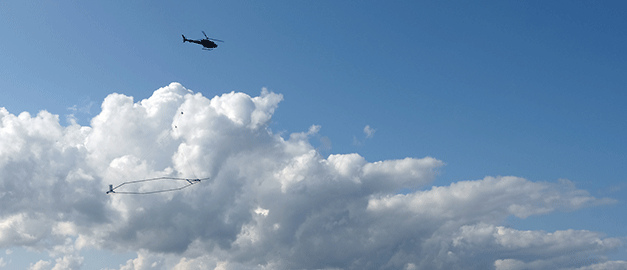 På bilden syns en helikopter som flyger. Under helikopter hänger ett stort mätinstrument. På bilden syns en helikopter som flyger. Under helikopter hänger ett stort mätinstrument.