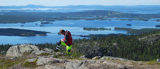 En man står och antecknar i ett block. I bakgrunden syns vatten och fjäll. Foto. En man står och antecknar i ett block. I bakgrunden syns vatten och fjäll. Foto.