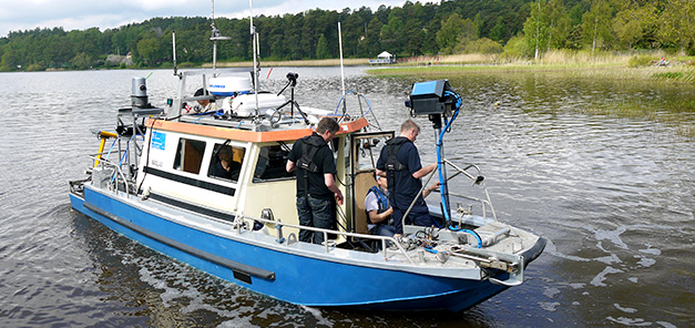 Tre personer arbetar på däck på en mindre blå och vit båt. Insjö med skog och strand i bakgrunden. Tre personer arbetar på däck på en mindre blå och vit båt. Insjö med skog och strand i bakgrunden.