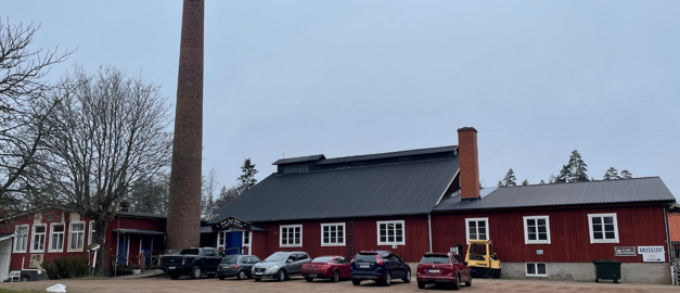 Bilden visar Bergdala Glasbruk och är tagen från parkeringsytan framför den rödmålade byggnaden. En hög skorsten sträcker sig upp mot himlen vid byggnadens ena sida.