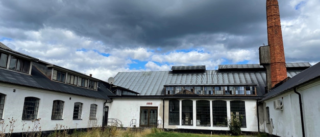 Bilden visar Flygsfors glasbruk med den visa fasaden och det svarta taket. En molning himmel syns bakom byggnaden.