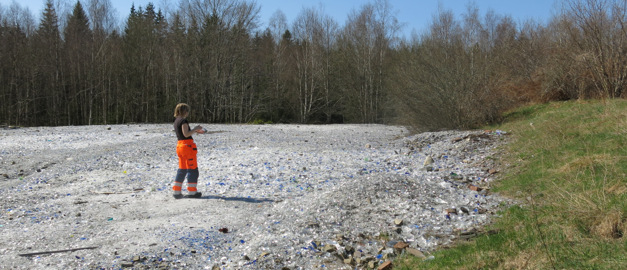 En blandning av klara och blåa glasskärvor breder ut sig över marken i den gamla glasdeponin. En kvinna står i glasfältet. En blandning av klara och blåa glasskärvor breder ut sig över marken i den gamla glasdeponin. En kvinna står i glasfältet.