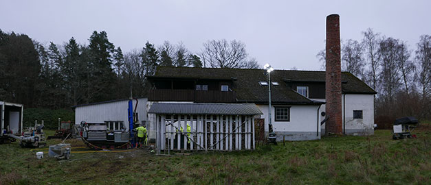 Person med varselväst och maskin sanerar marken bredvid ett hus med tegelskorsten.